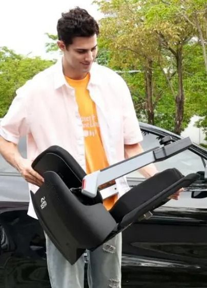 Man holding a black folded car seat by a black car, smiling, outdoors.