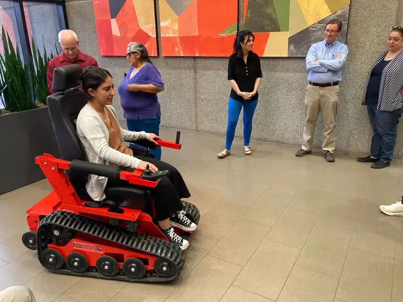 Woman in a red tracked wheelchair demonstrates it to a group of people indoors, looking pleased.