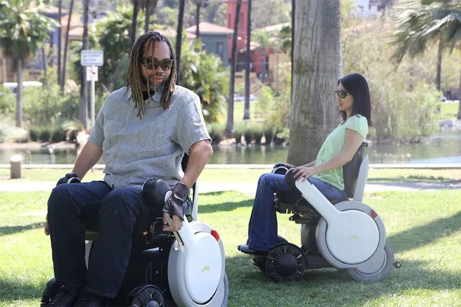 Two people in power wheelchairs outdoors near a lake.