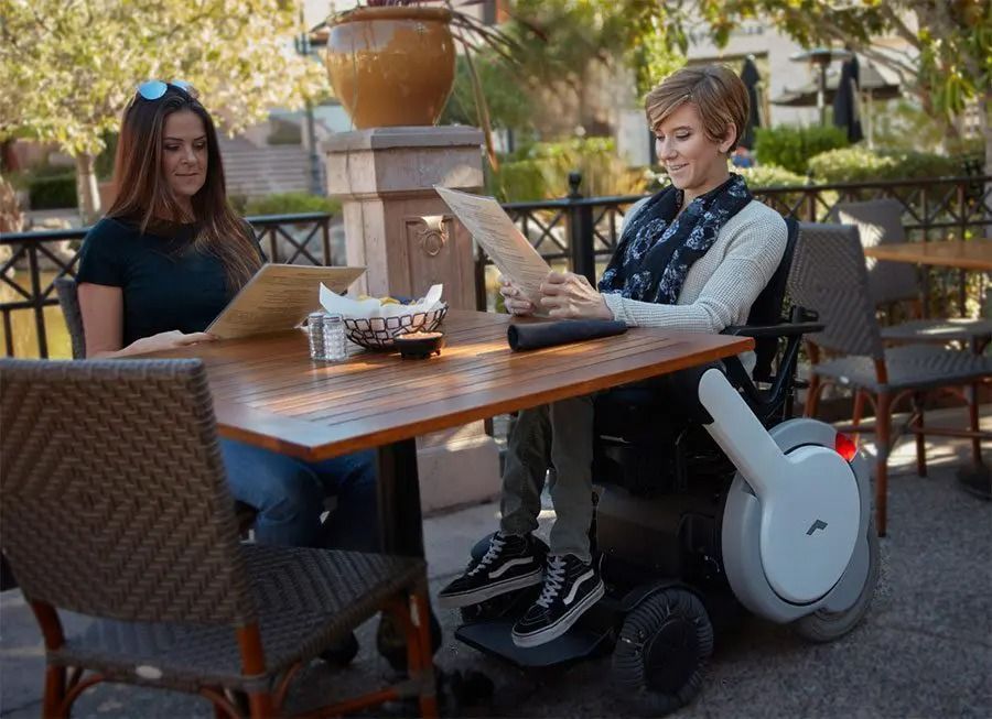 Two women at a cafe table, one in a wheelchair, looking at menus outdoors.
