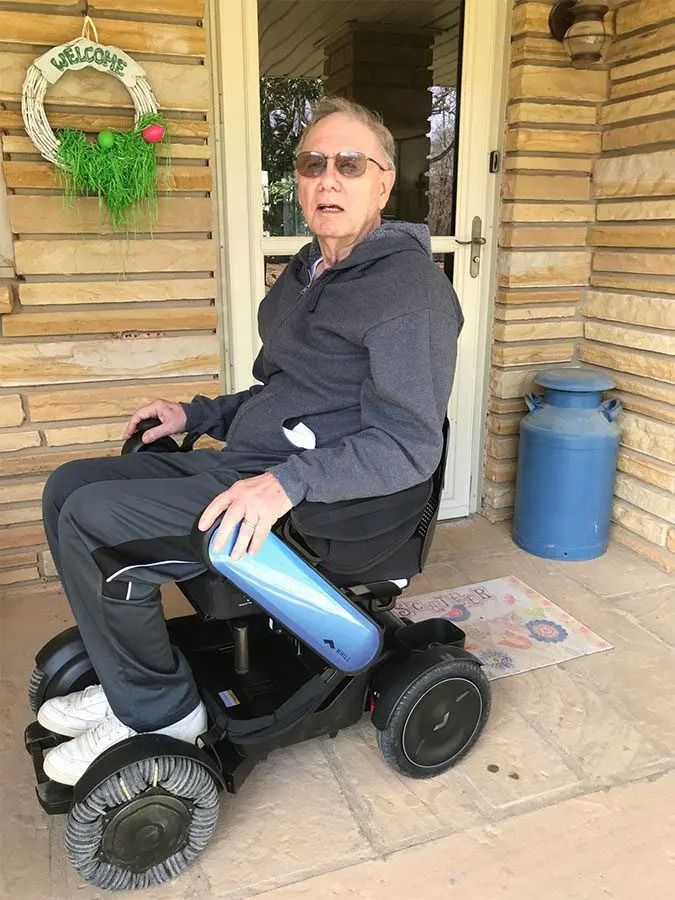 Man in a mobility scooter on a porch, wearing gray clothing, with a blue container in the background.