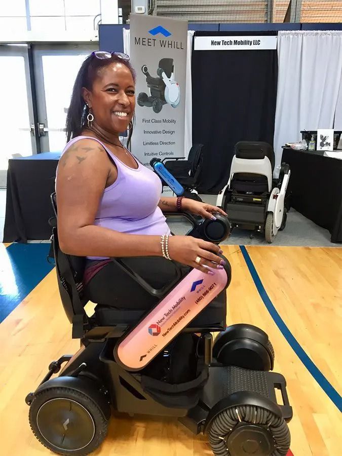 Woman in a power wheelchair smiles in a venue. Purple tank top, black hair, wood floor, two other wheelchairs.
