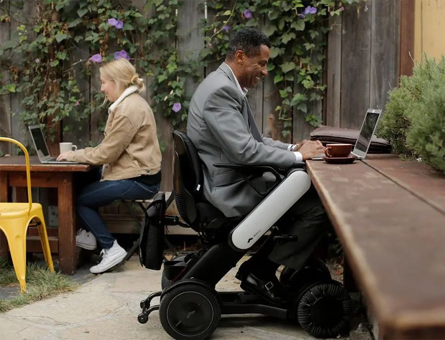 A man in a wheelchair and a woman work on laptops outdoors at a shared table.