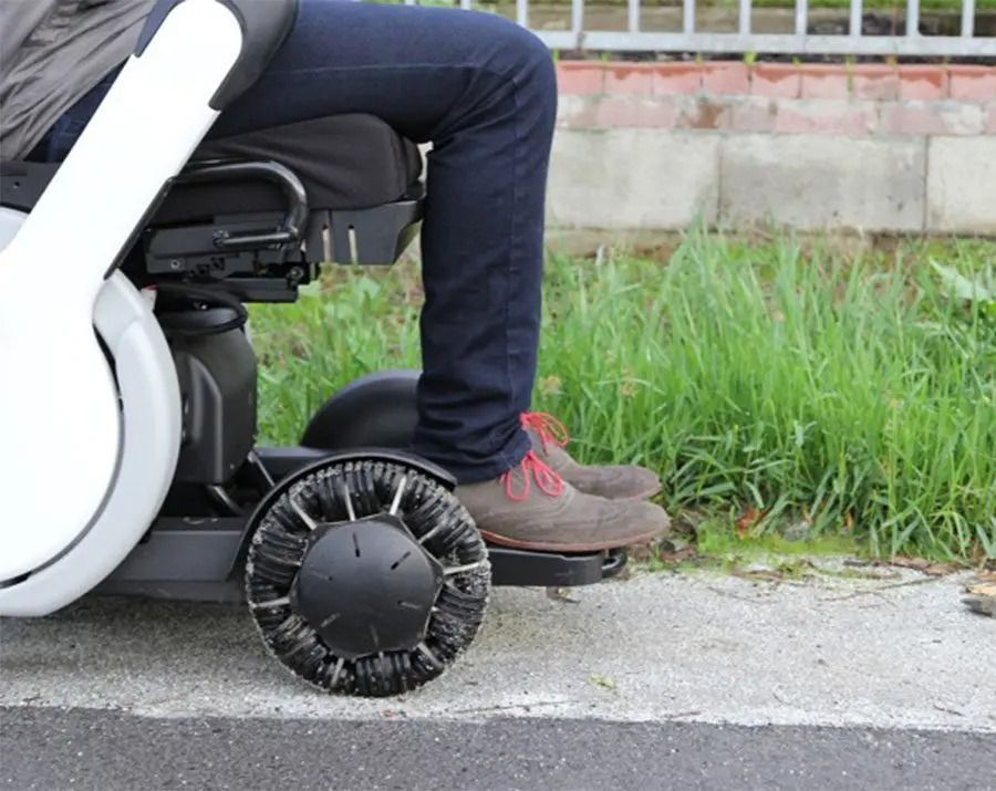 Person in a wheelchair, next to a curb, with the wheel's black tread contrasting with the sidewalk.