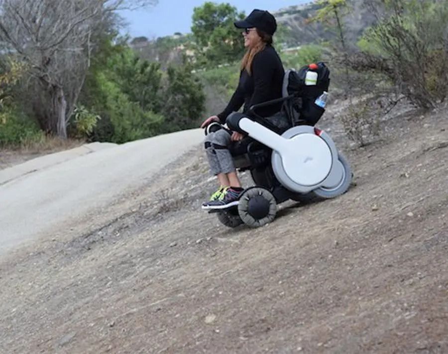 Woman in black outfit riding power wheelchair up a dirt path.