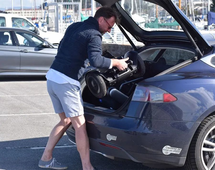 Man in shorts loads a black mobility scooter into the trunk of a blue car, near a harbor.