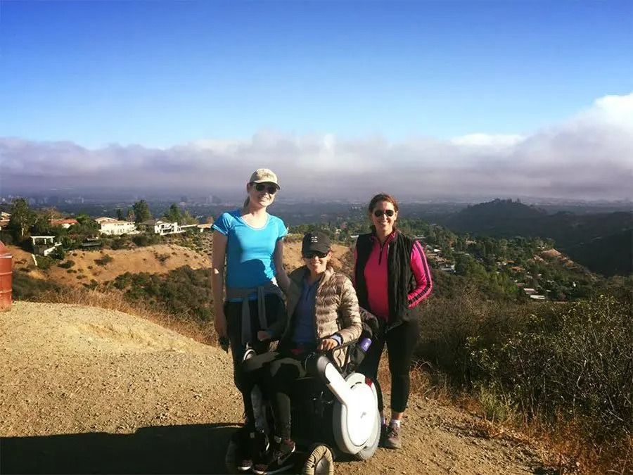 Three women on a hilltop. One in a wheelchair smiles, flanked by standing women. Cloudy valley background.
