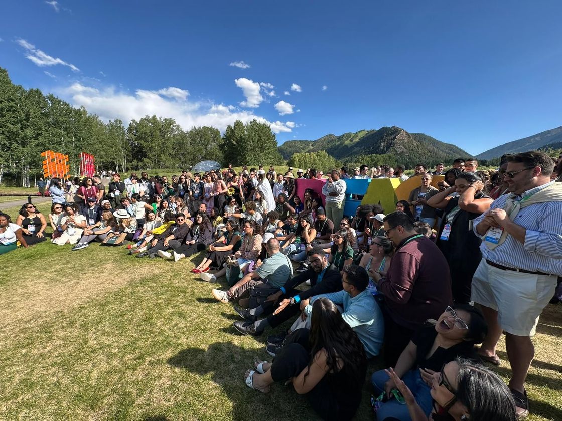 Crowd seated on grass at an outdoor event under a bright blue sky with trees and a hill in the background