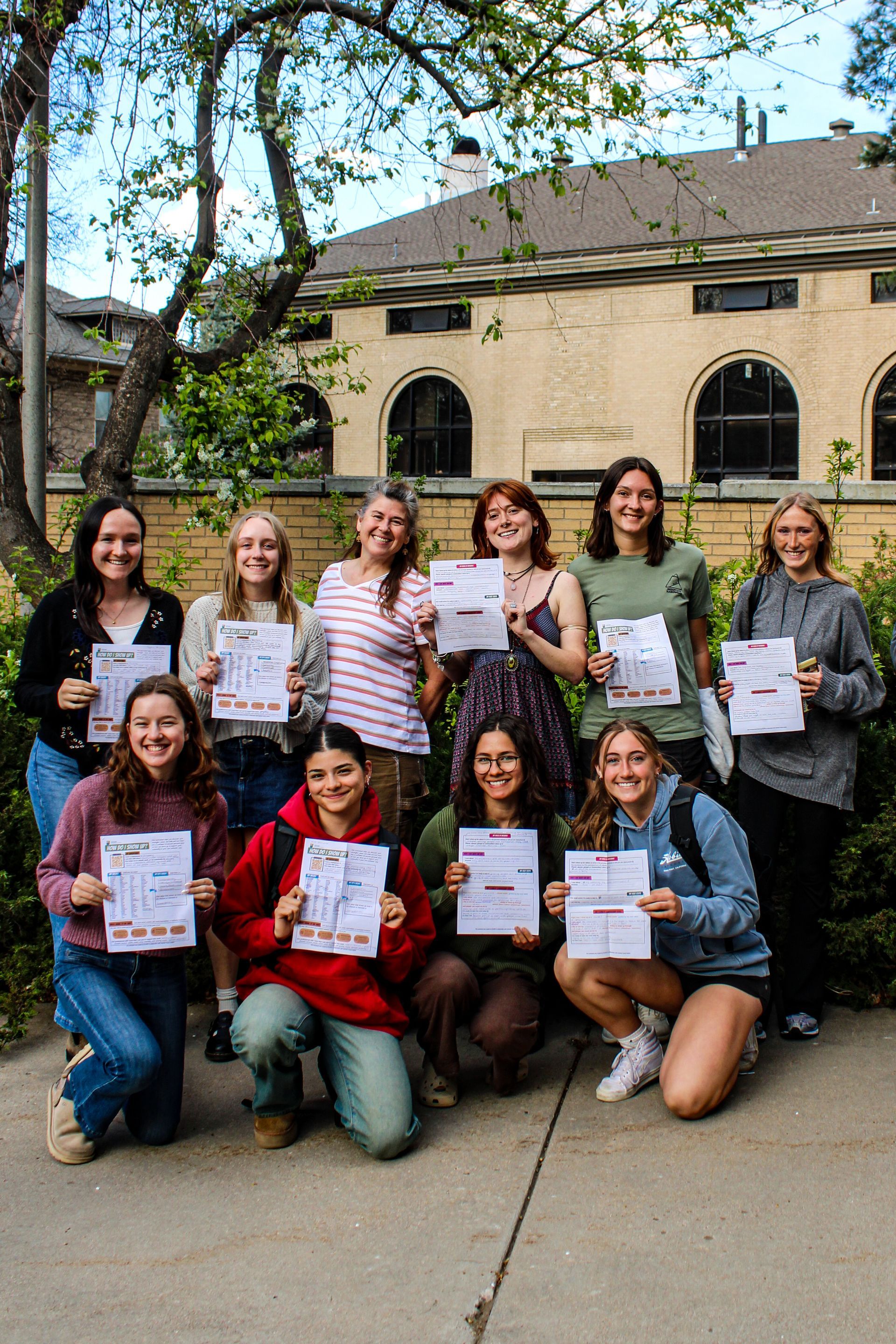 A group of twelve people smile while standing outdoors, each holding up a flyer in front of a tan building.