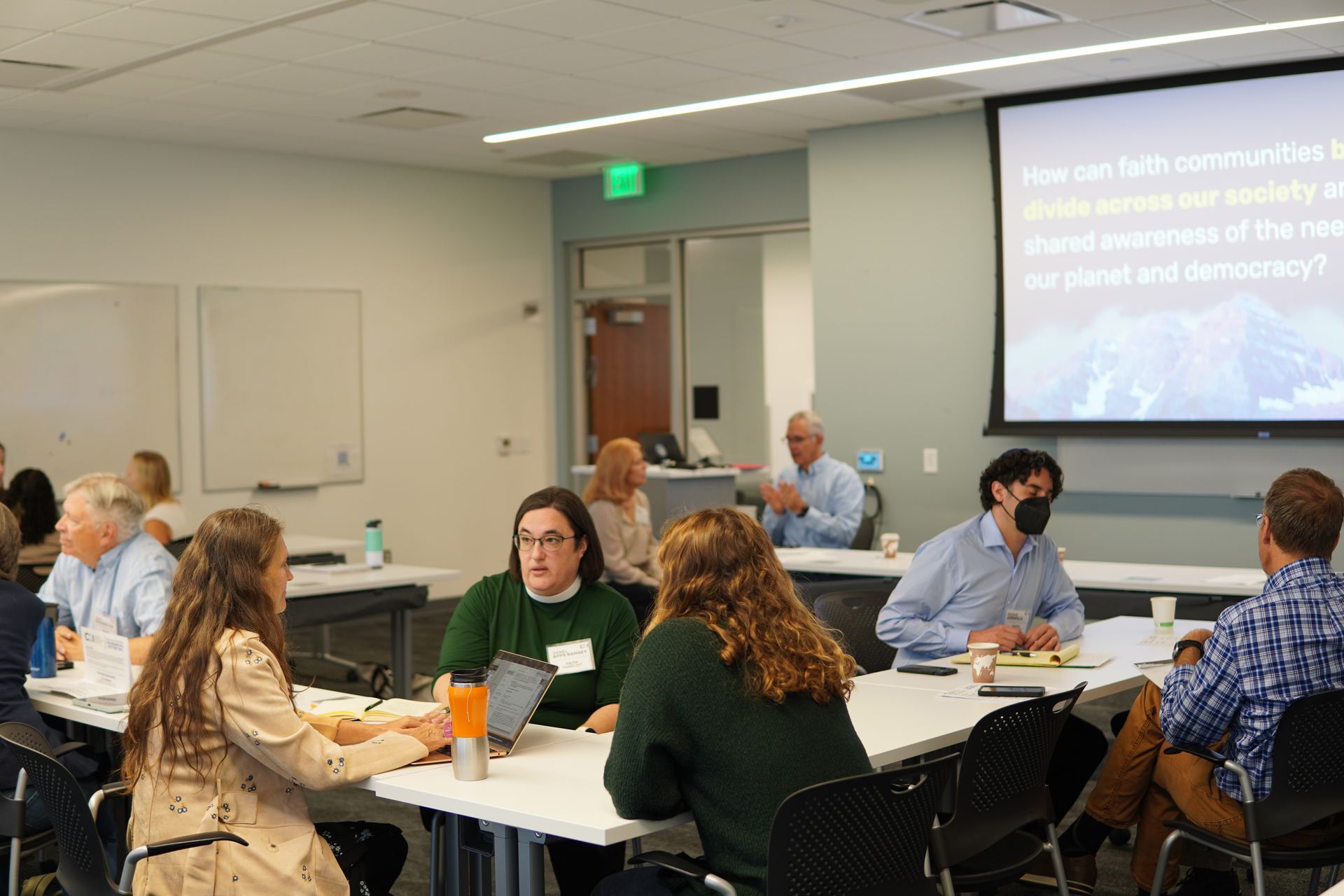 People sit at tables in a brightly lit room with a presentation screen discussing the health of the planet and democracy.