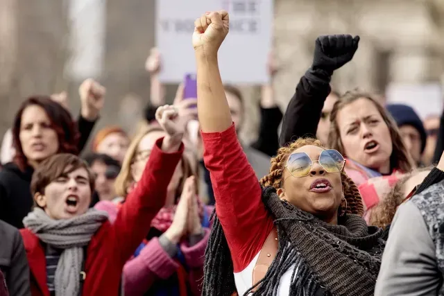 A crowd of protesters shouting, with several people raising their fists in the air at a public demonstration.