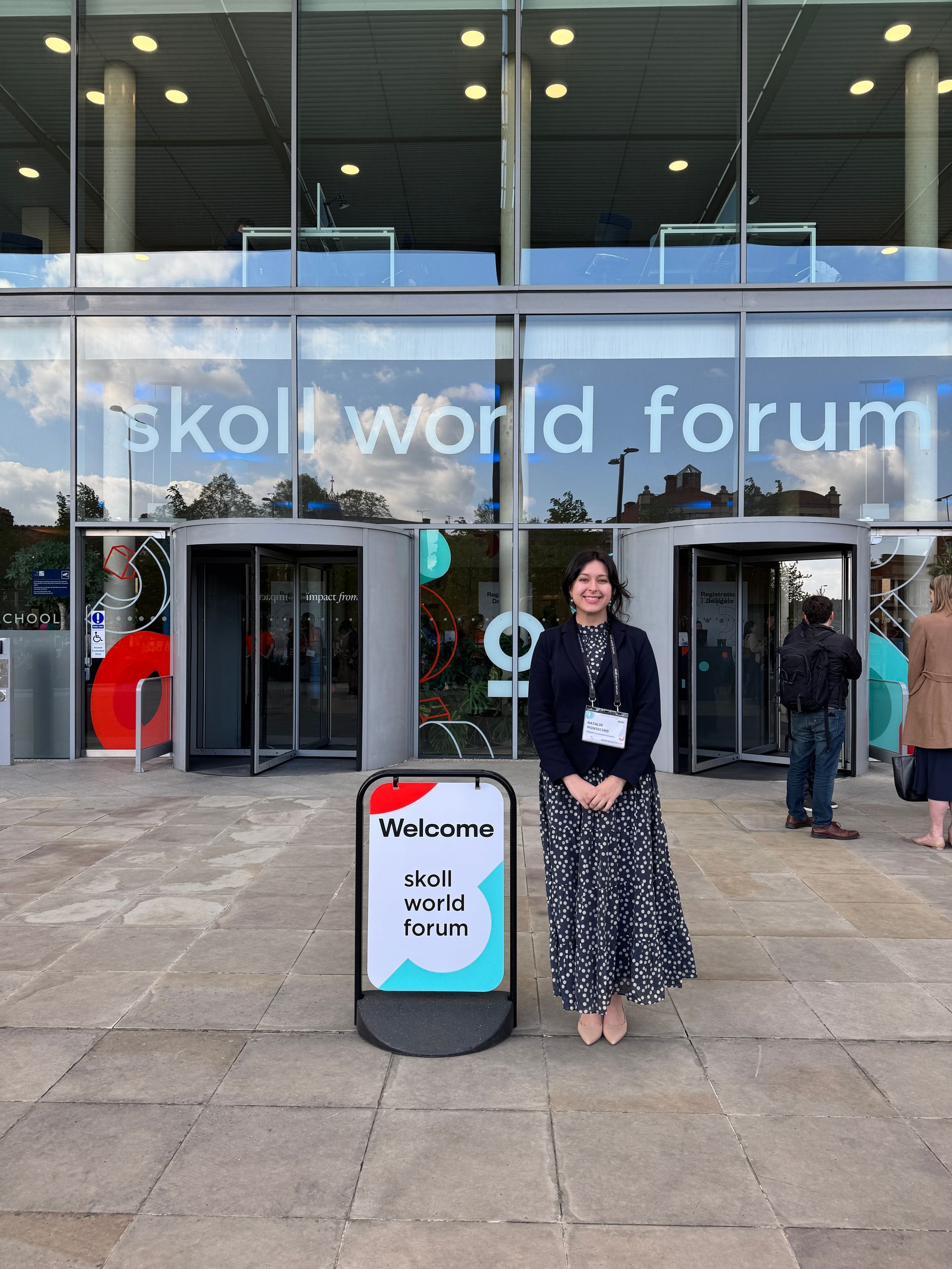 Crowd seated on grass at an outdoor event under a bright blue sky with trees and a hill in the background