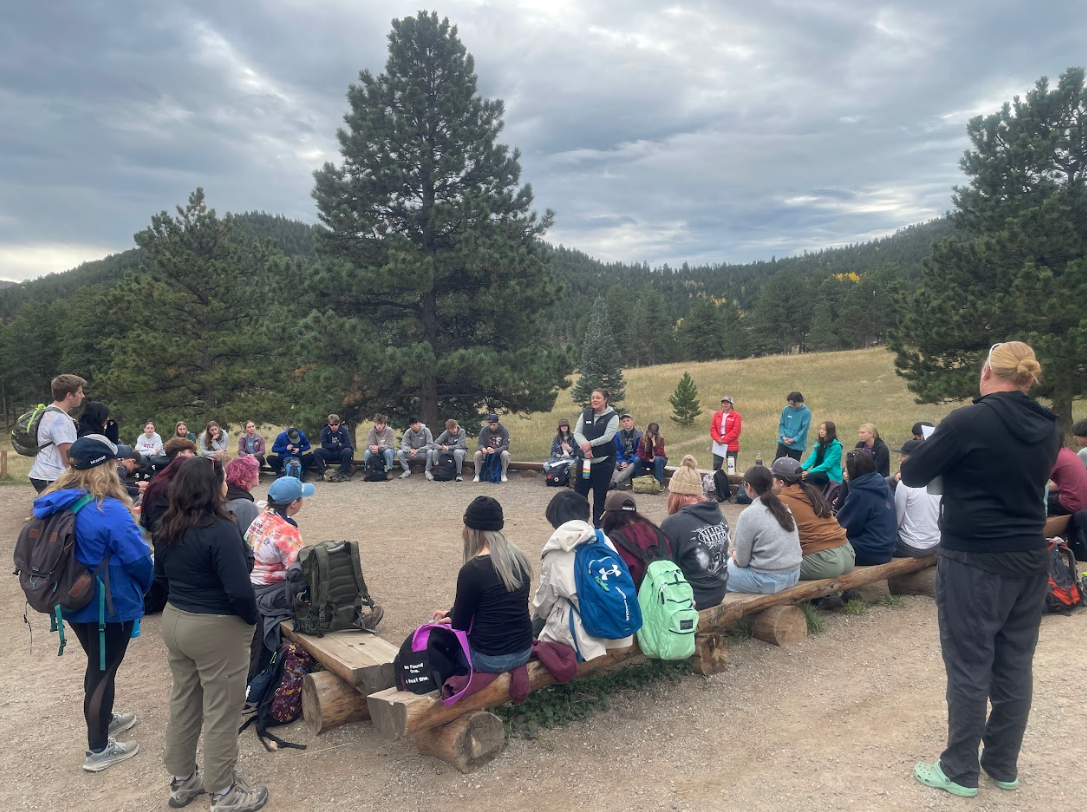Group seated on logs around a campfire in a forest clearing, with one person speaking nearby
