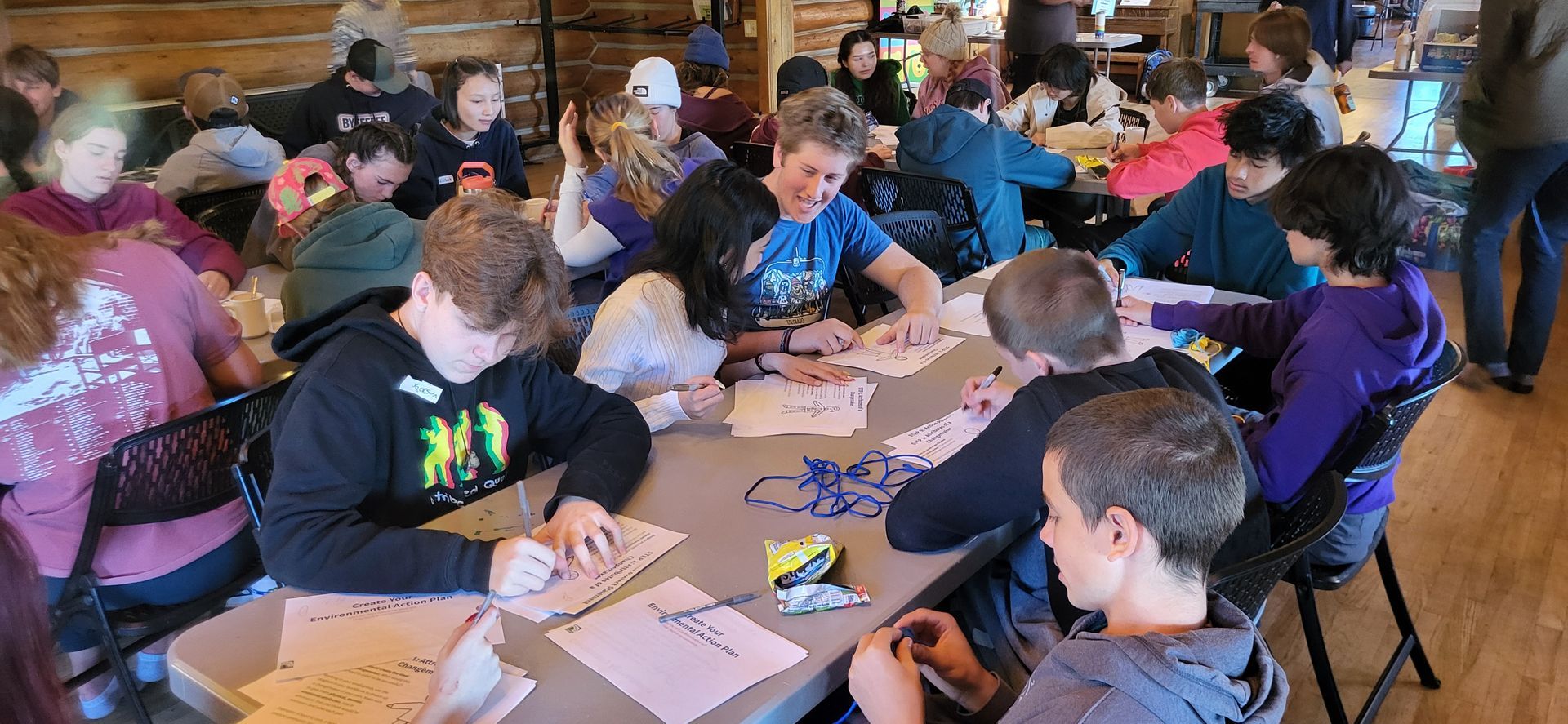 A group of people sits at long tables in a room with wood walls, working on papers and craft projects together.