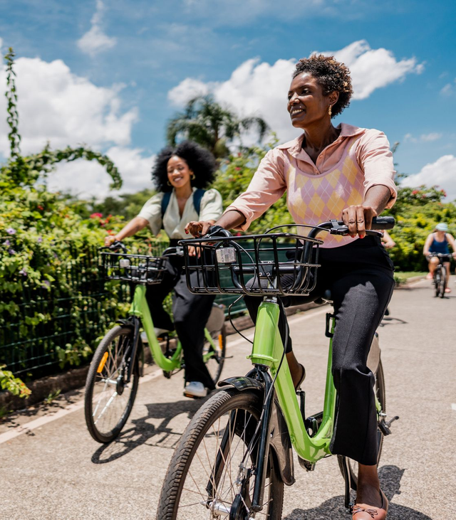 Two people riding bicycles on a sunny path, with front baskets and greenery along the trail.