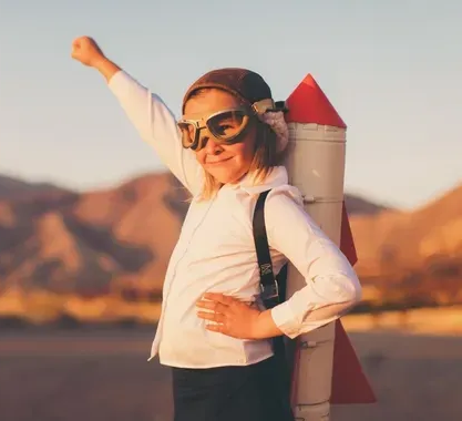 A child in a white shirt and aviator goggles stands with one arm raised, wearing a toy cardboard rocket on their back.