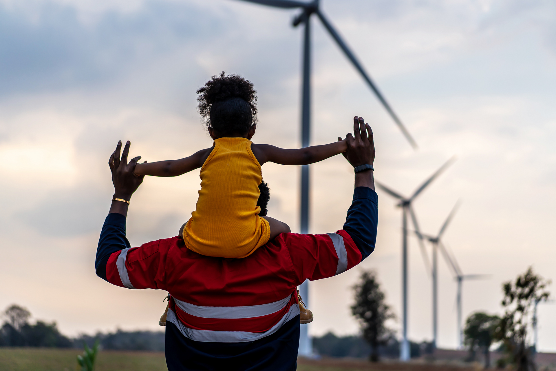 A person in a red and white work shirt carries a child in a yellow shirt on their shoulders against a wind farm background.