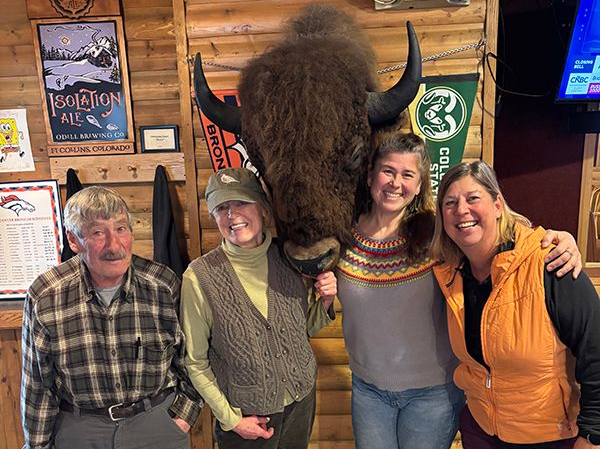 Four smiling people posing indoors beside a large mounted bison head in a rustic wooden room