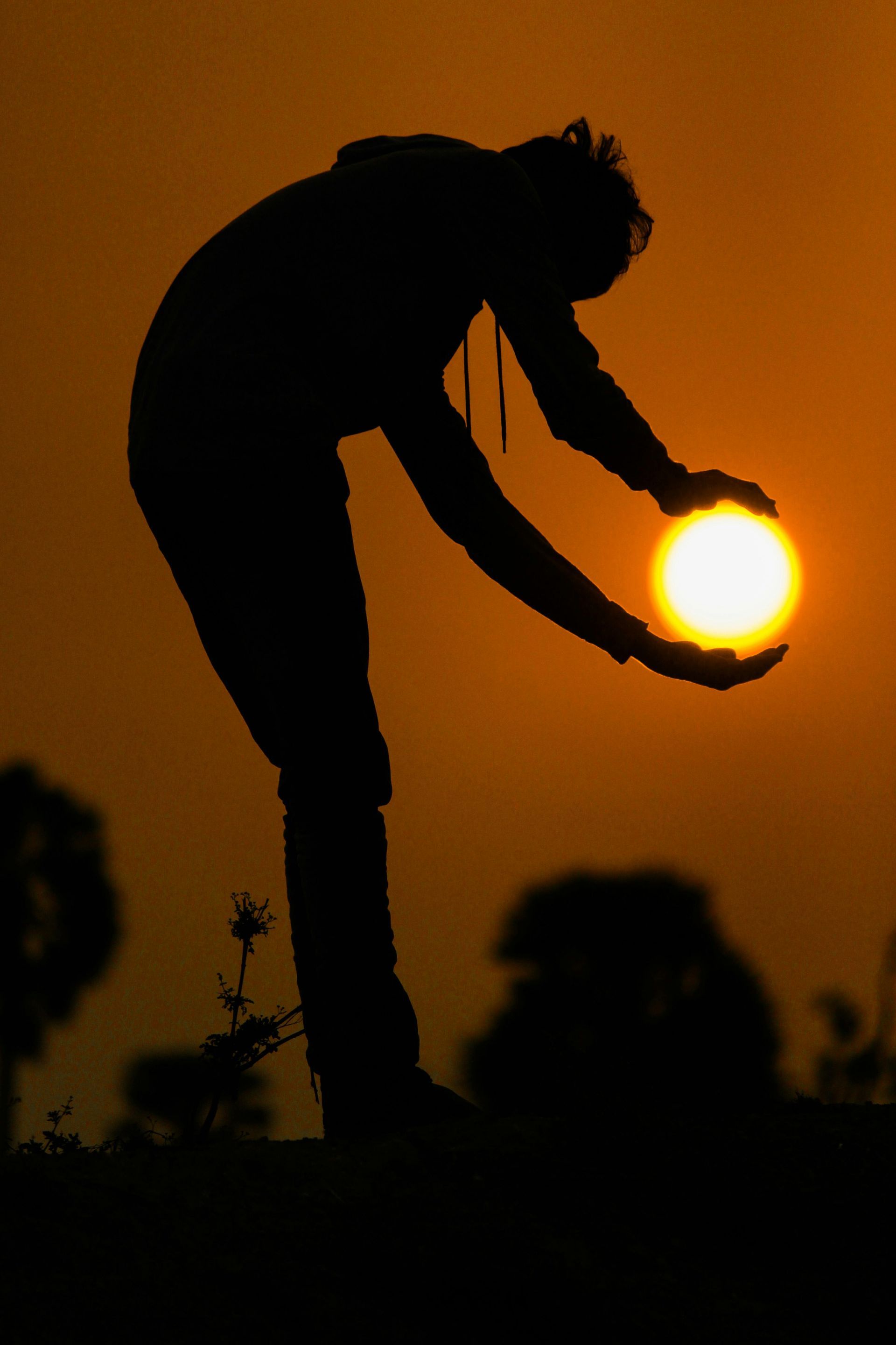 A silhouette of a person posing as if holding the glowing sun between their hands against an orange sky.