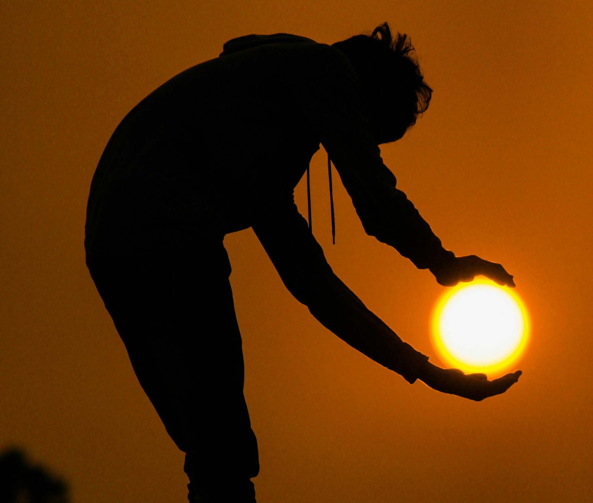 A silhouette of a person holding the setting sun between their cupped hands against an orange sky.