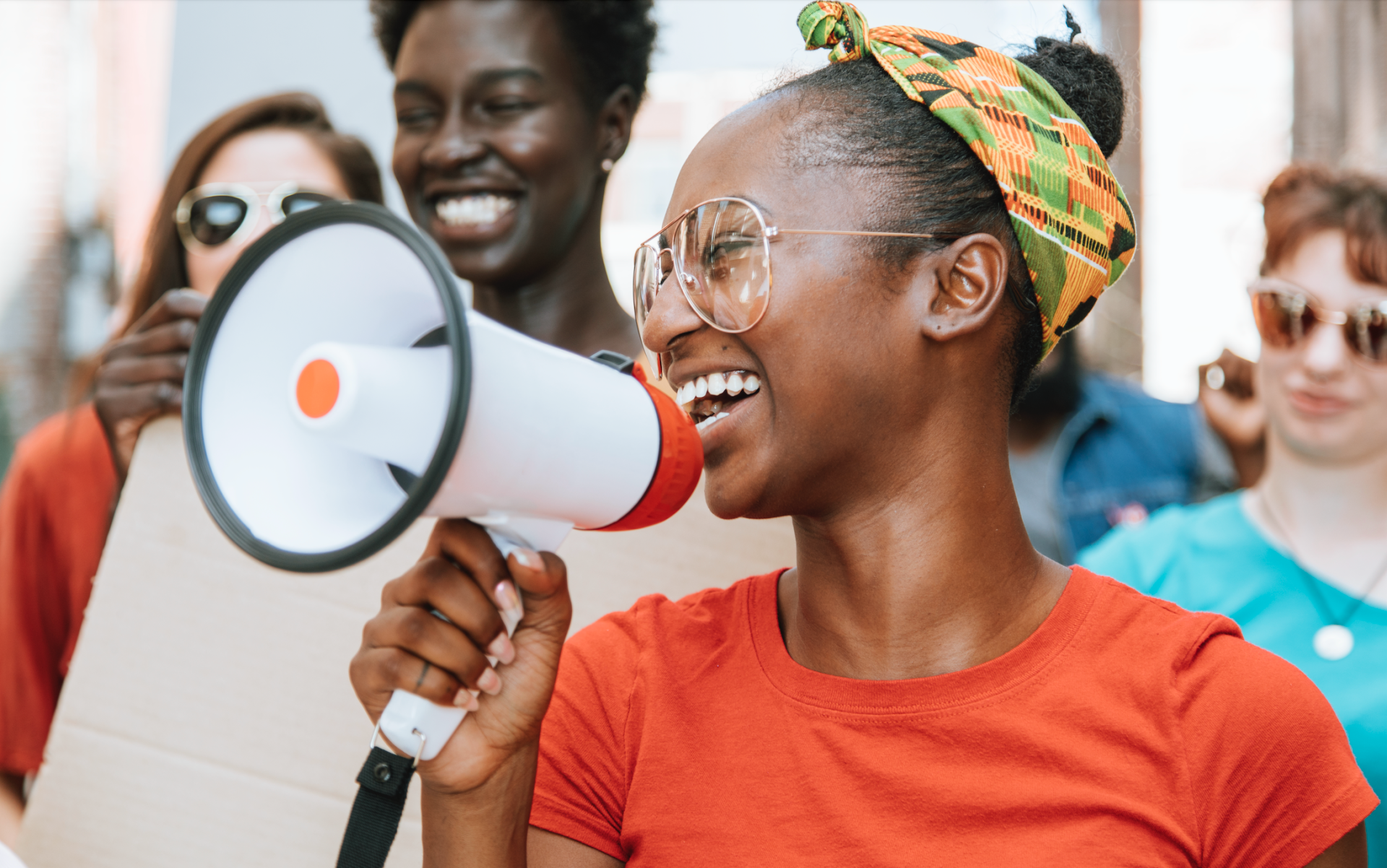 A person wearing glasses and a colorful headband smiles while shouting into a white megaphone during a protest.
