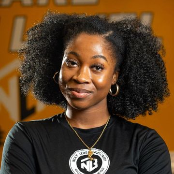 A woman with curly hair is wearing a black shirt and a necklace. Coach Alexa