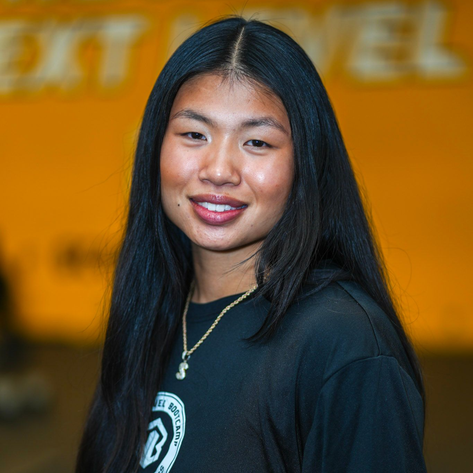 A woman with curly hair is wearing a black shirt and a necklace. Coach Alexa