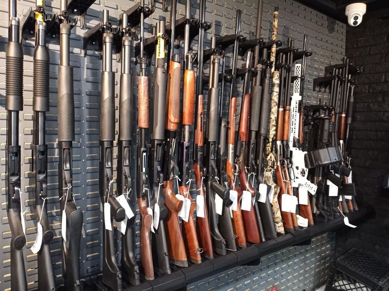 A variety of shotguns and rifles hanging vertically on a pegboard wall display inside a retail store.