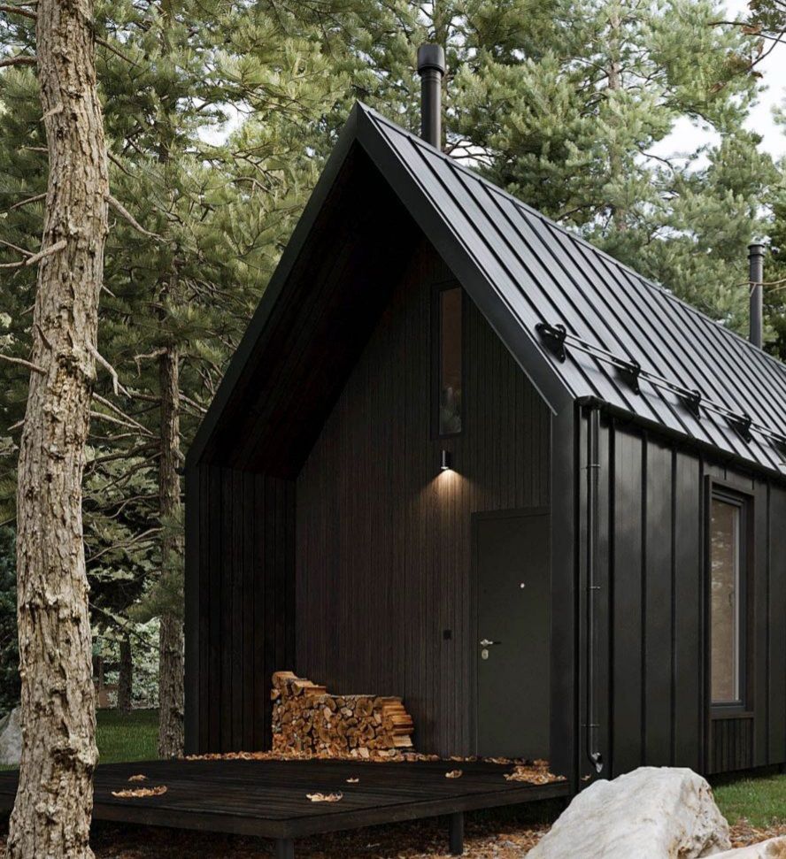 Black cabin in a forest, with woodpile on the porch, dark siding and metal roof.