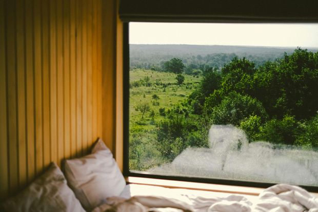 Bed beside a window overlooking a green landscape with trees, wooden walls, and white pillows.