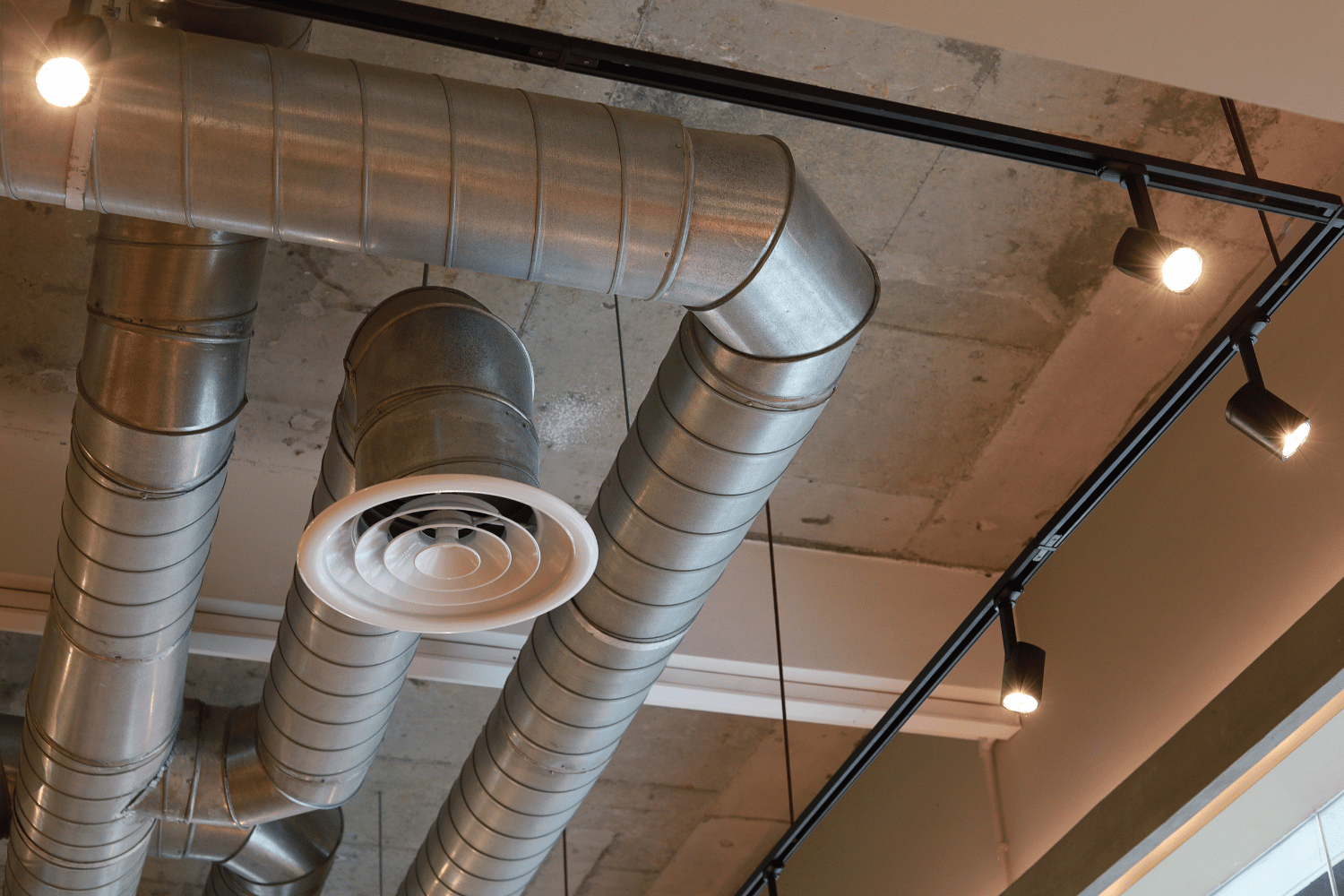 Exposed ceiling with silver ventilation ducts, a round white air vent, and track lighting on a textured concrete surface.
