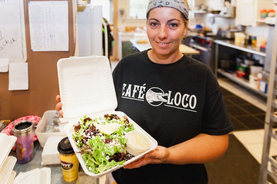 A woman wearing a cafe loco shirt is holding a styrofoam container of food.