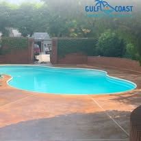 A swimming pool with turquoise water, surrounded by a brown patio, in front of a building and bushes.