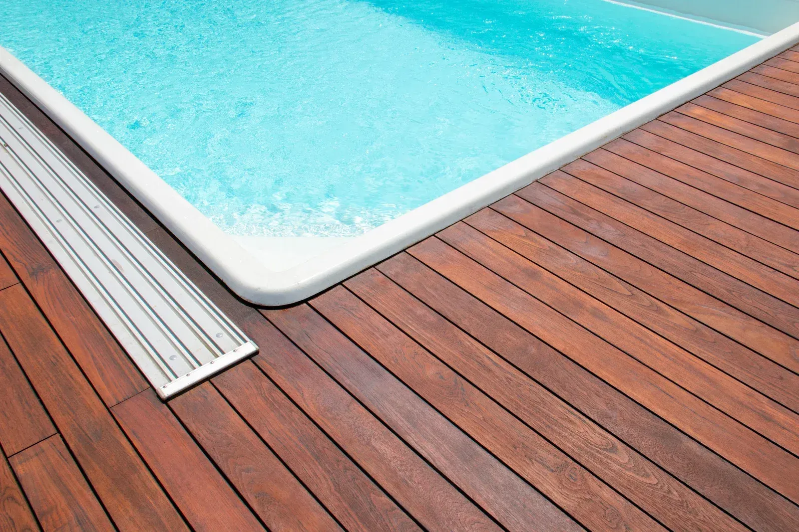 Wooden deck bordering a turquoise pool; a metallic drainage strip separates the deck from the pool's edge.