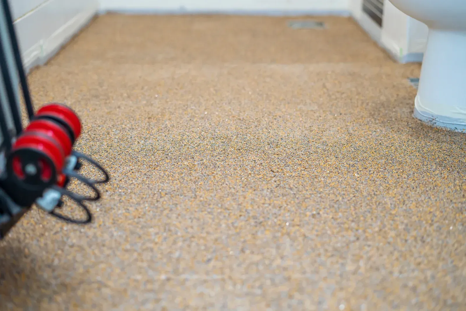 Beige, textured flooring in a bathroom; a toilet and a red object with black tubing are visible.