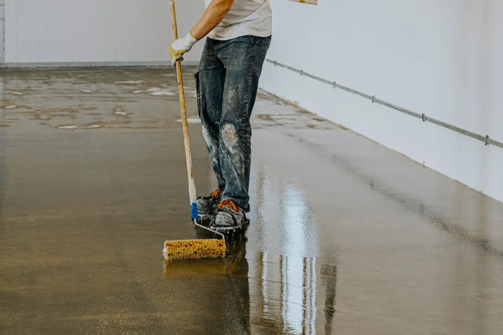 Person applying sealant to a concrete floor with a roller in a white-walled room.
