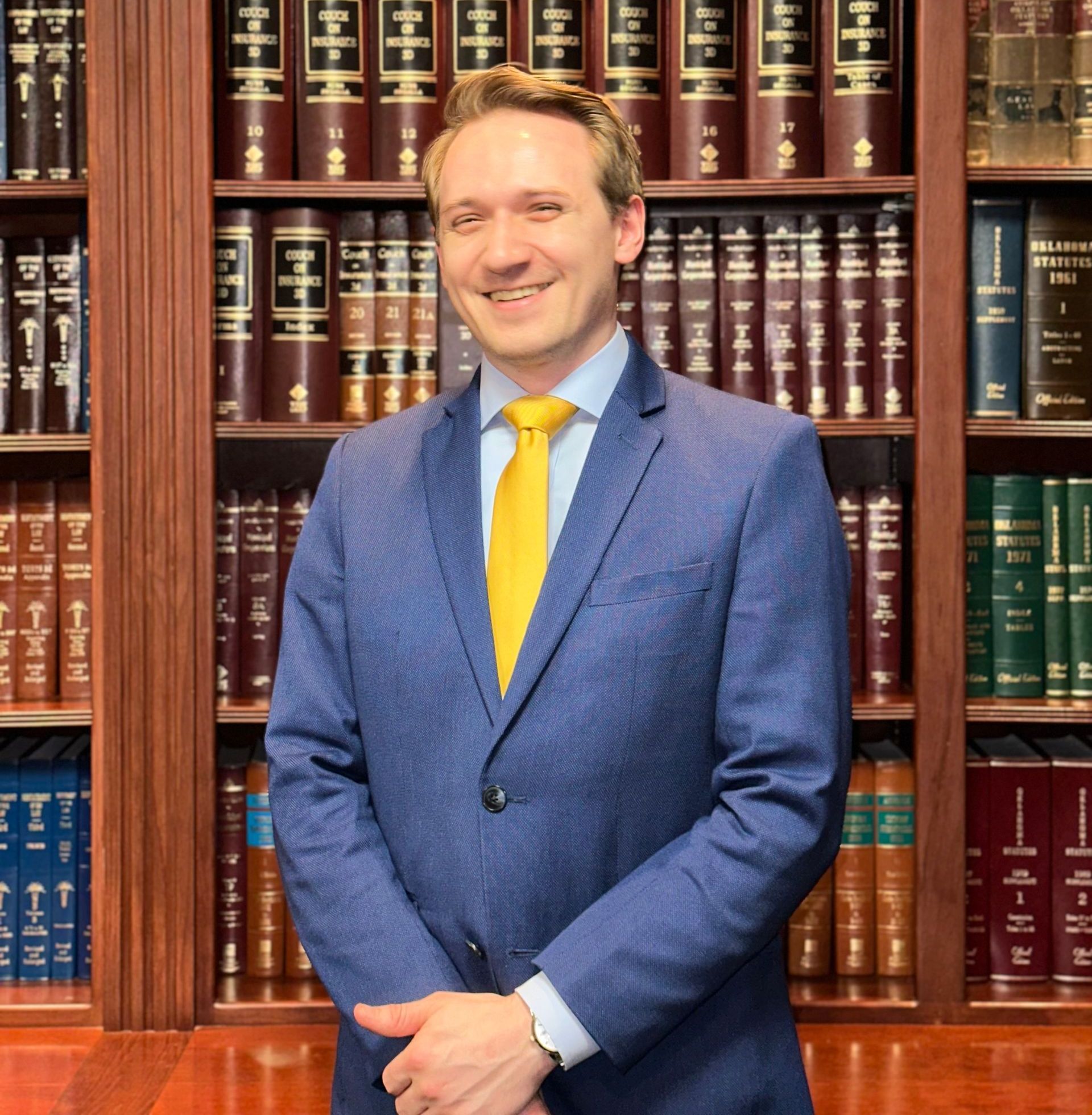 A man in a blue suit and yellow tie stands in front of a bookshelf
