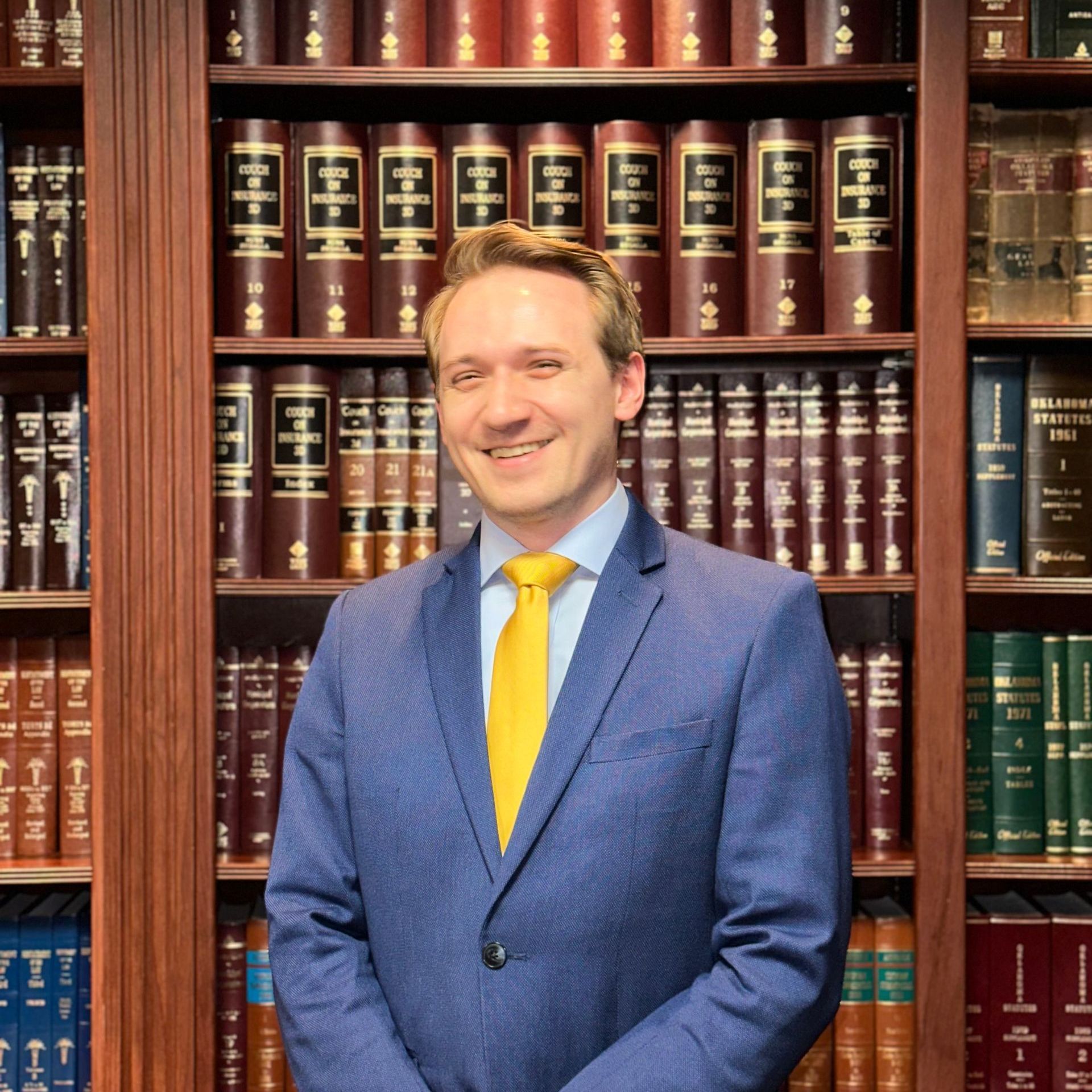 A man in a blue suit and yellow tie stands in front of a bookshelf full of books