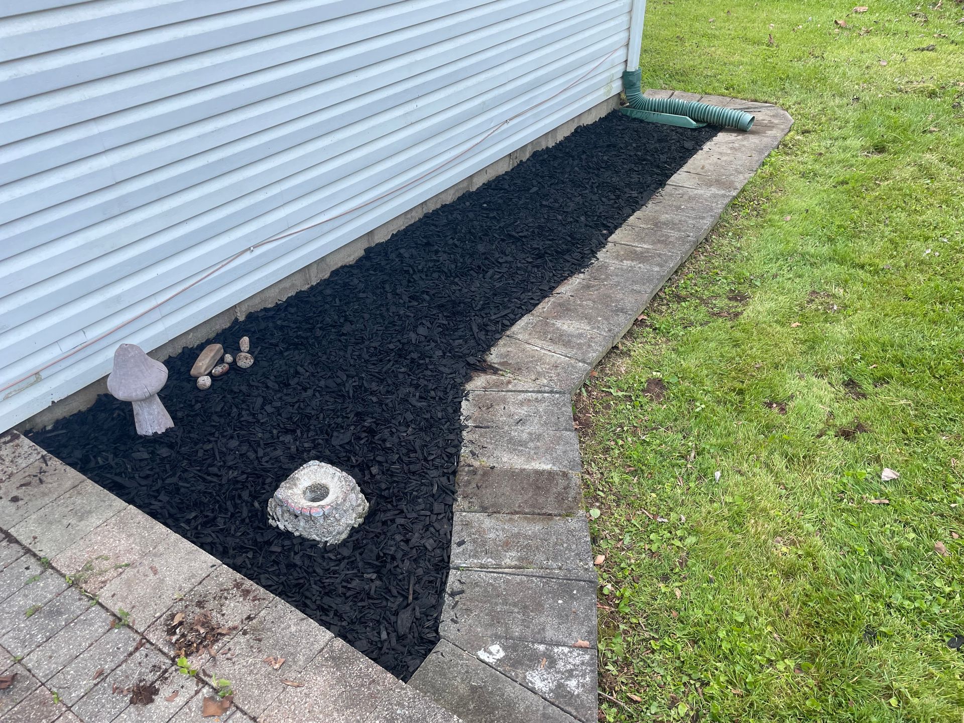Mulched garden bed beside a white building, bordered by pavers and lawn, with small ornaments.