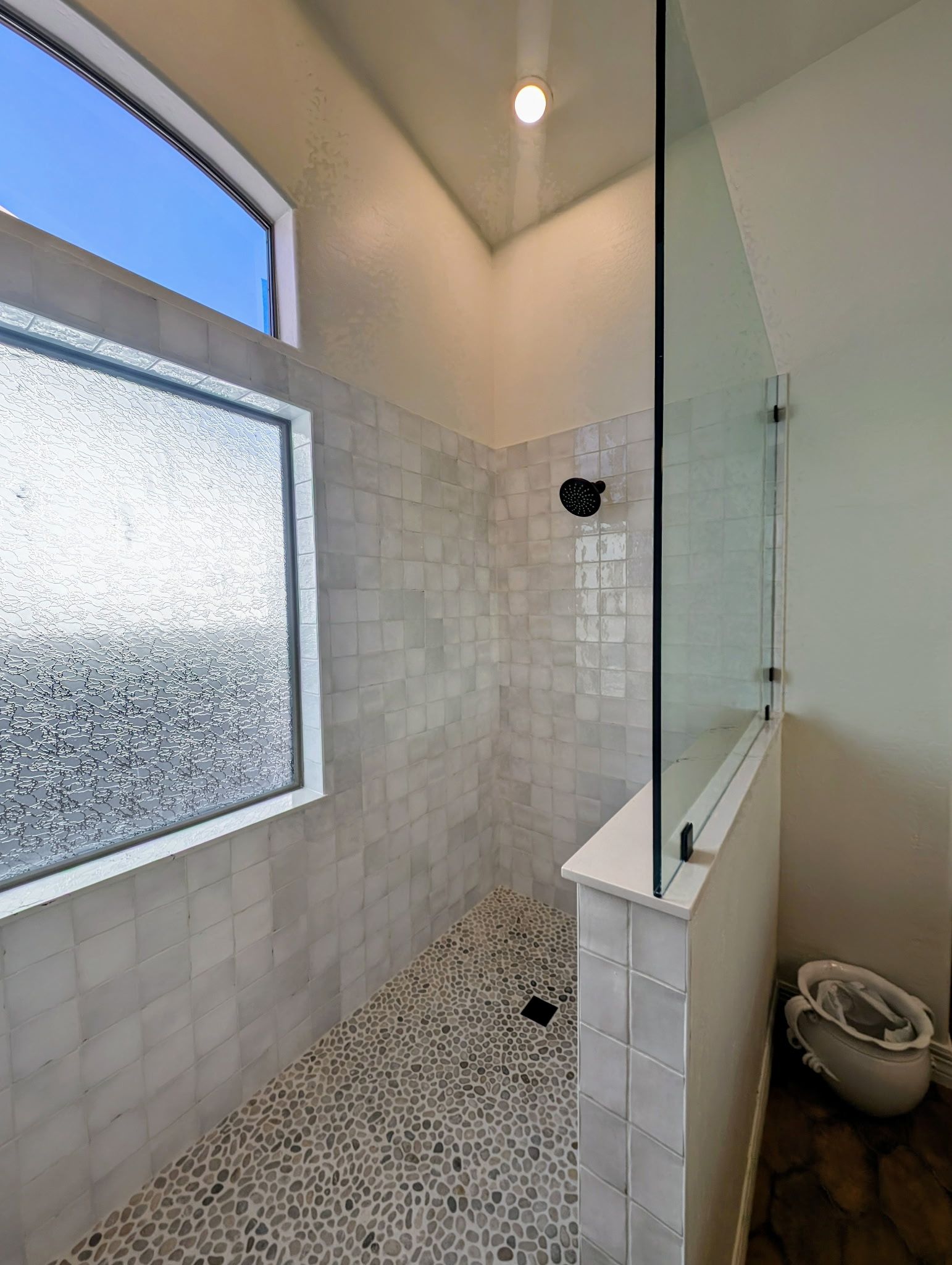 Shower with pebble floor, glass door, and frosted window. Light-colored tiles and walls.