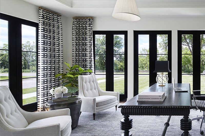 A living room with a desk , chairs , and windows.
