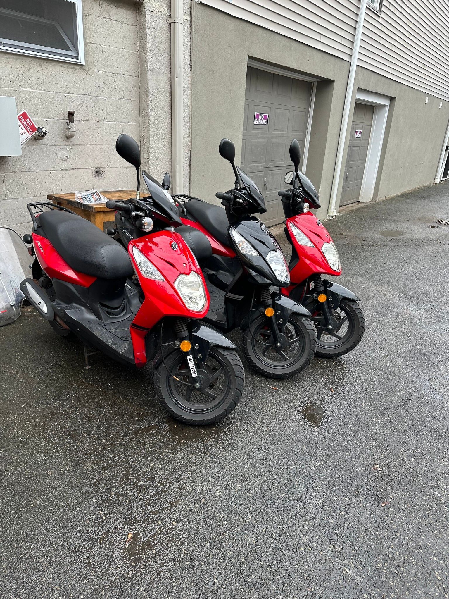 Three red and black scooters parked on a wet, paved surface next to a building.