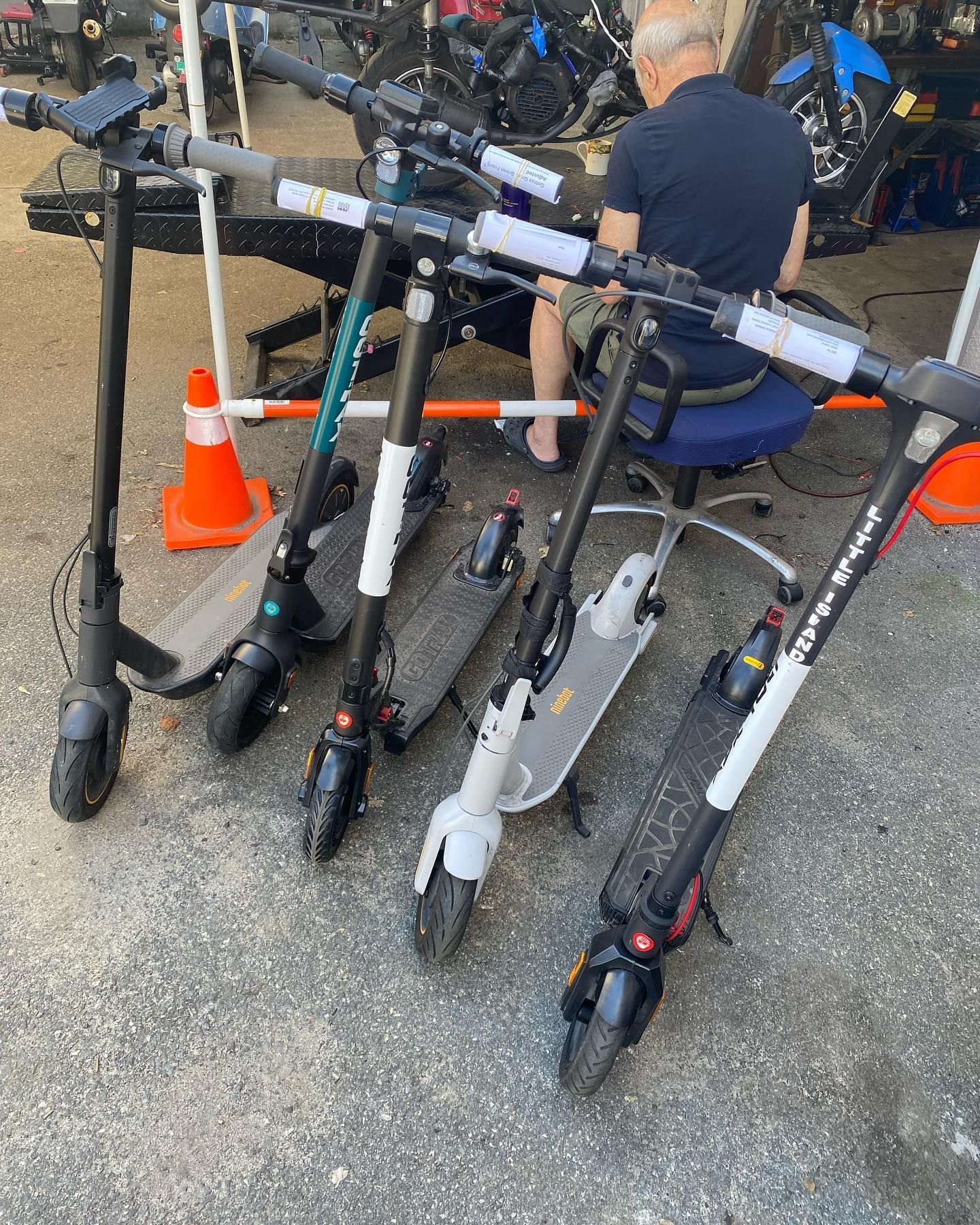 Electric scooters parked on pavement, person seated nearby, cones present.