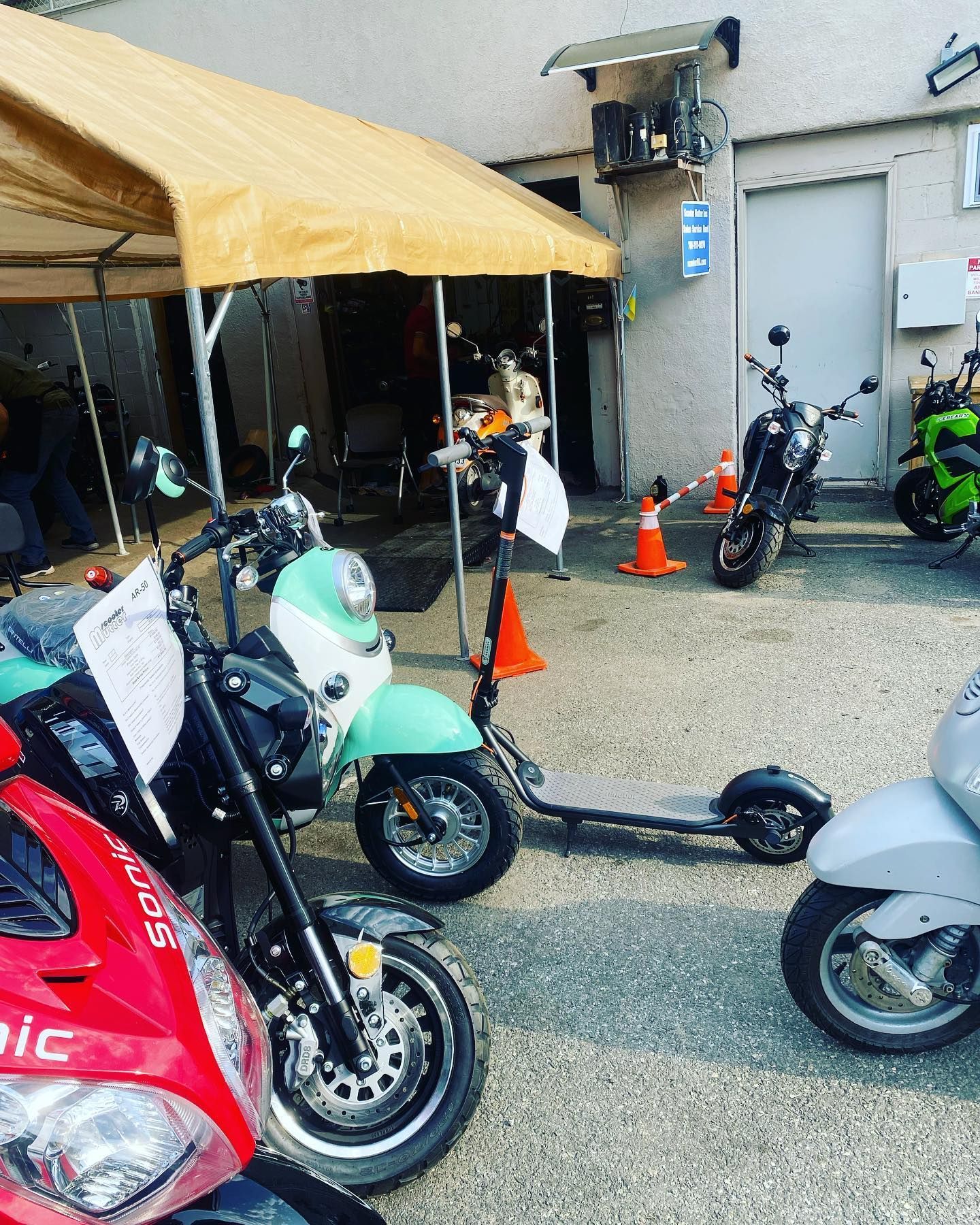 Motor scooters of various colors parked outside a building with a canopy.