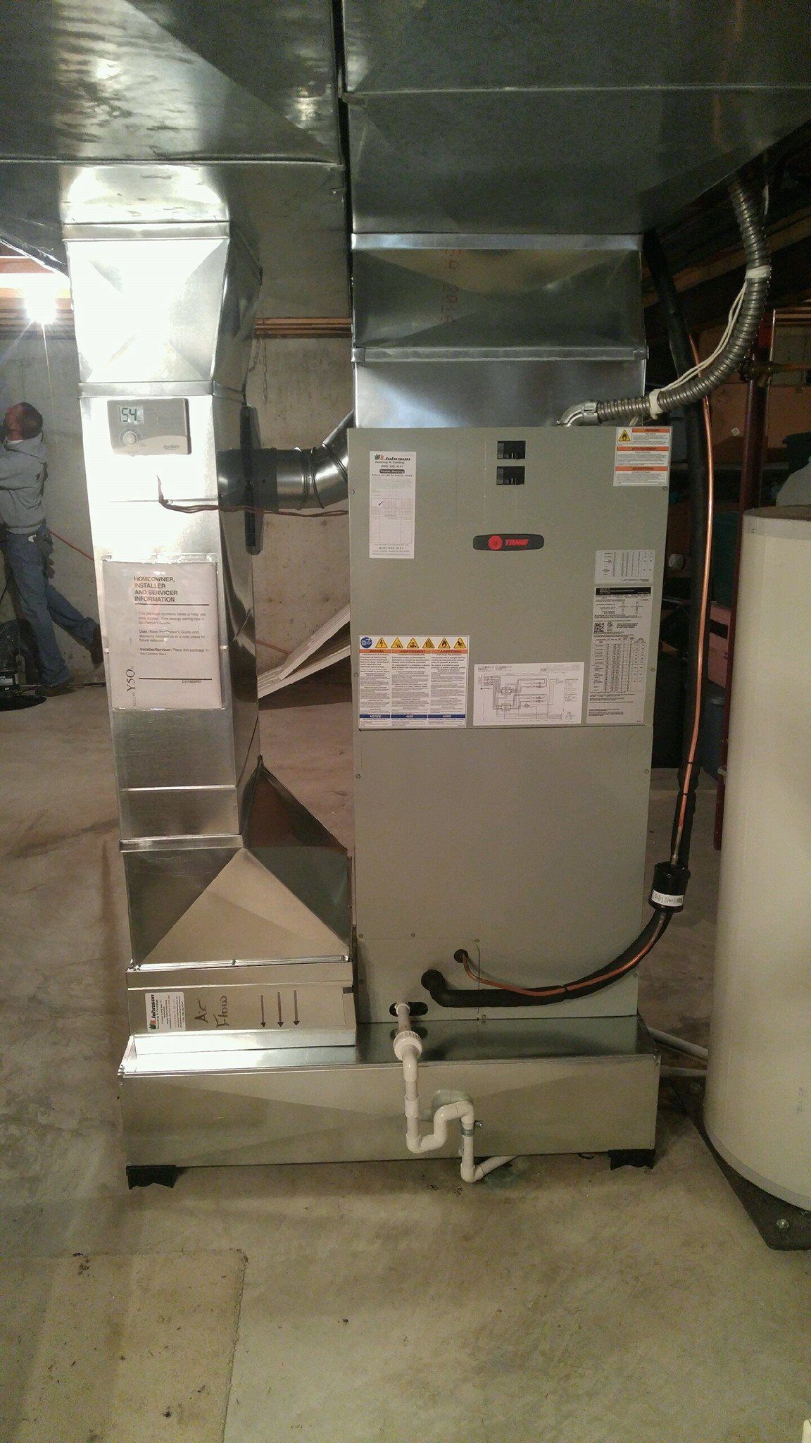 A large air conditioner is sitting in a basement next to a water heater.