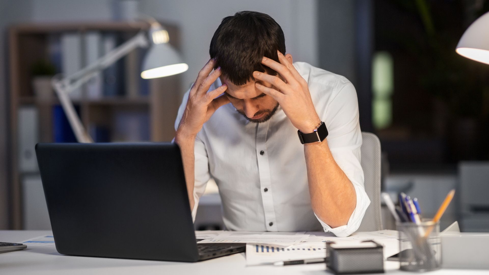 A stressed person sitting at a desk in an office with their head in their hands while working on a laptop.