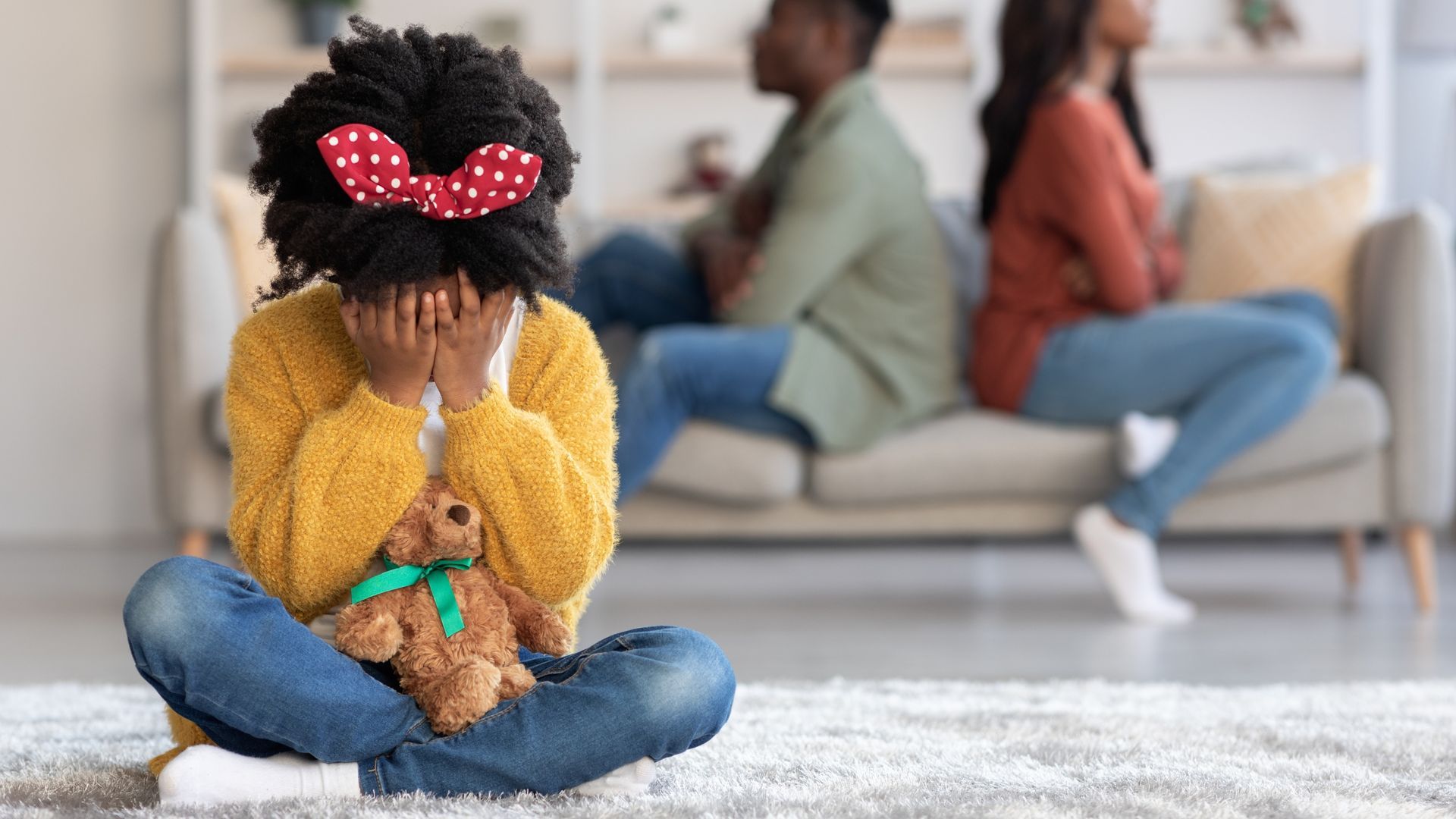 A child sits on the floor clutching a teddy bear with their hands over their face, while two adults sit apart on a sofa.