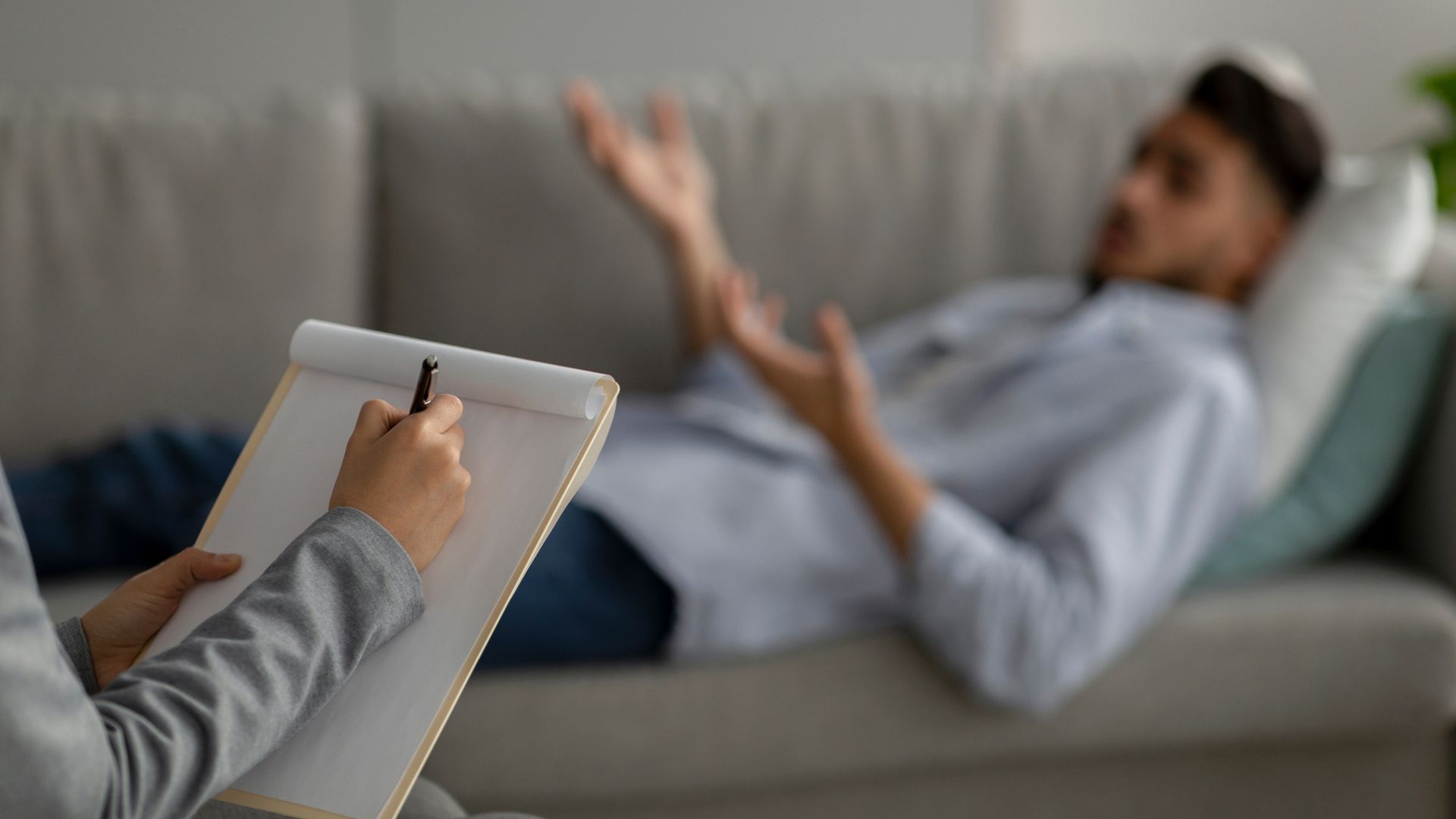 A person takes notes on a clipboard while a patient lies on a sofa and gestures during a therapy session.