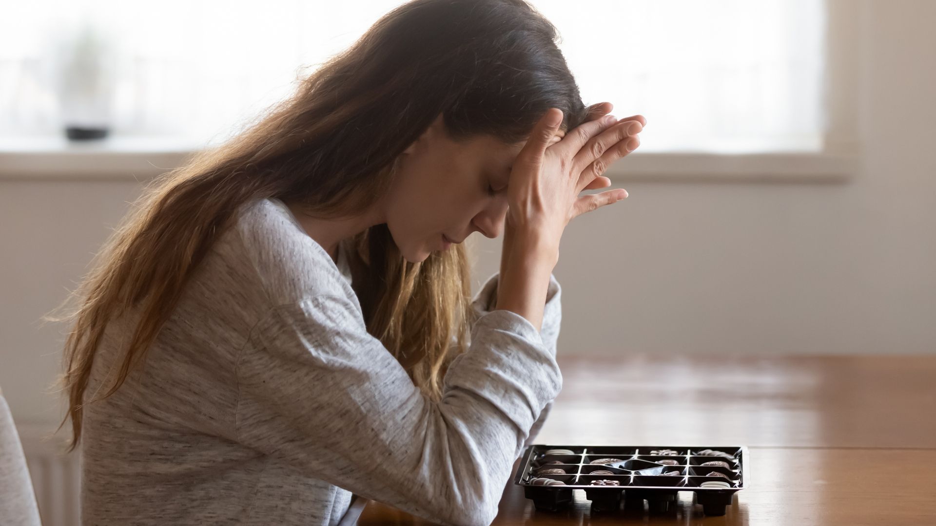 A person sits at a wooden table with their head in their hands, looking down at an empty plastic chocolate tray.