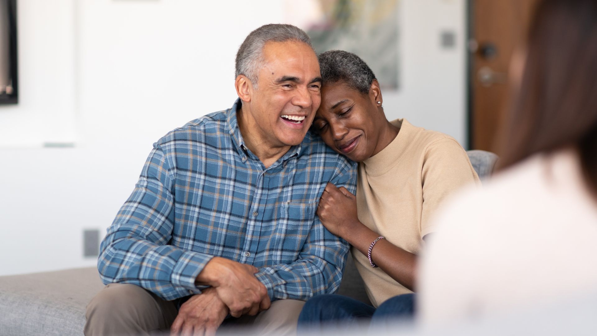 A couple sitting on a couch, with one person leaning their head on the other's shoulder while both smile in conversation.